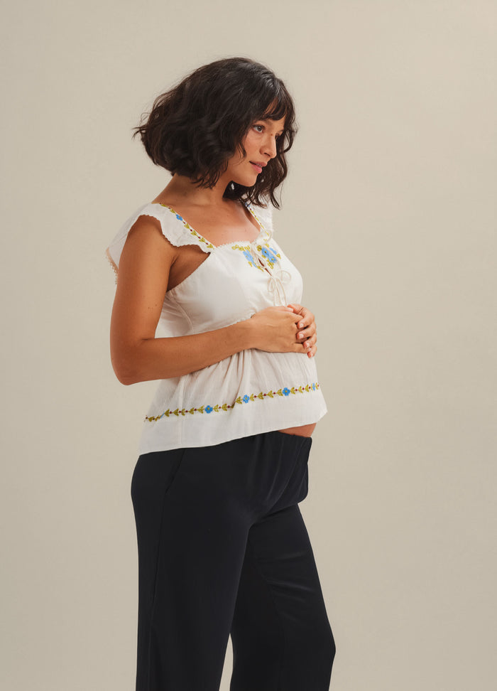 White sleeveless maternity top with ruffled straps and colorful embroidered trim across chest and hem.