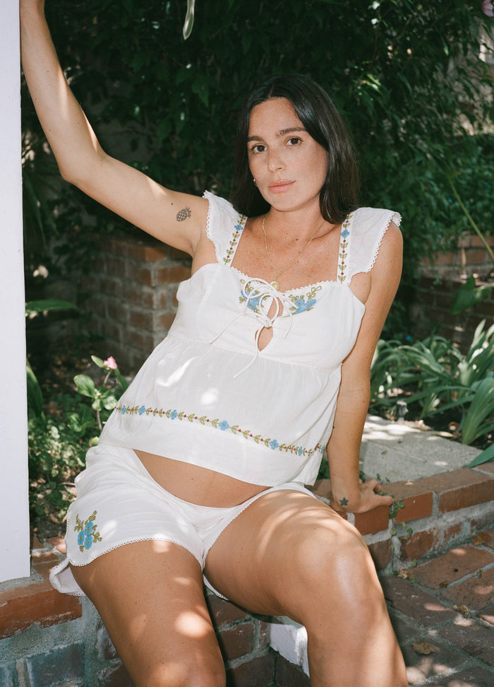 White sleeveless maternity top with ruffled straps, square neckline, and floral embroidery detailing.