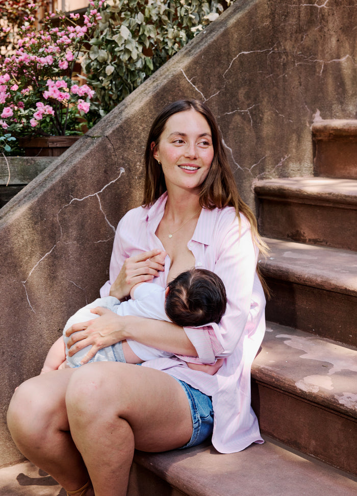 Light pink maternity buttondown shirt with long sleeves, worn open over white nursing bra, paired with denim shorts.