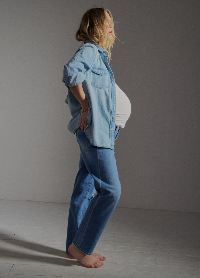The Denim Maternity Shirt - Light wash denim maternity shirt with rolled sleeves, worn open over a white tank top during pregnancy.