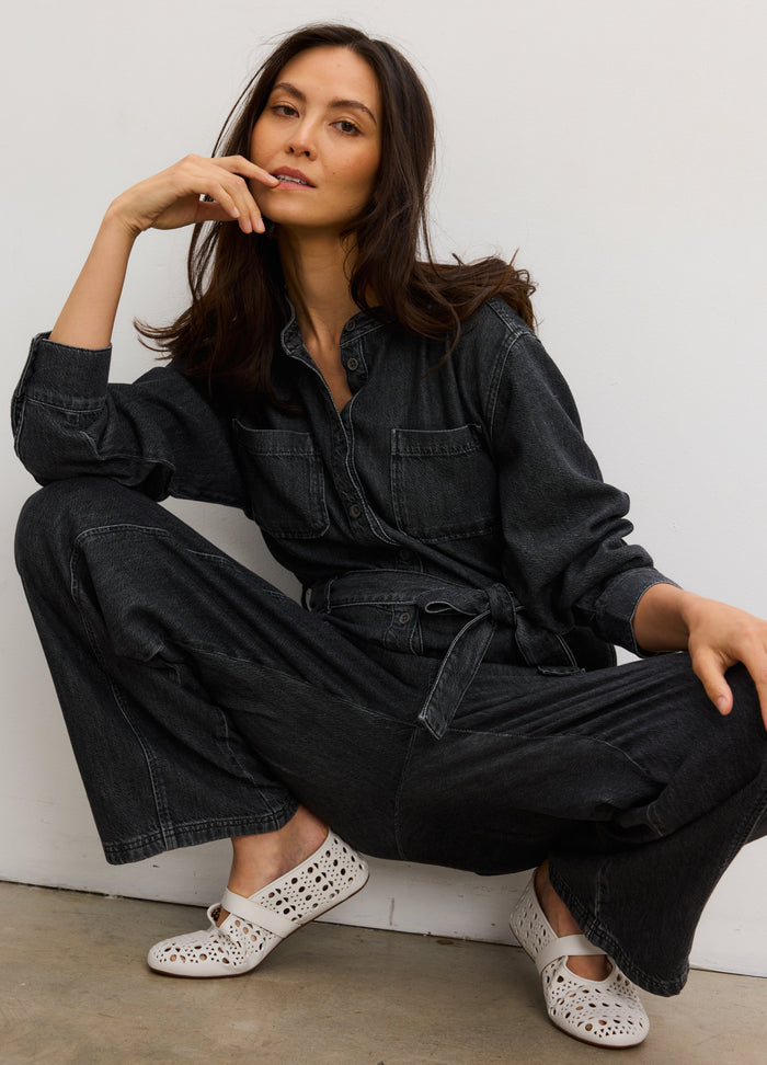 Pregnant woman wearing washed black drapey denim maternity jumpsuit with button-up front and belted waist.