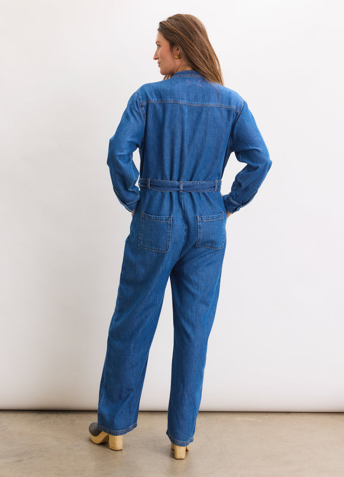 Blue denim maternity jumpsuit with long sleeves, gathered waist, and relaxed fit from the back.