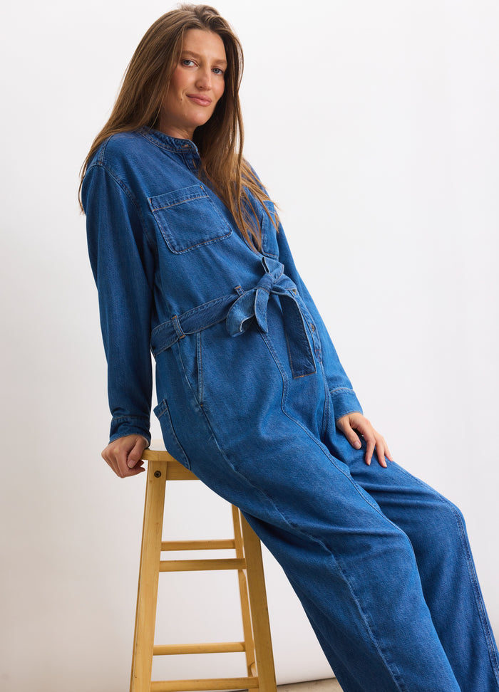 Blue denim maternity jumpsuit with long sleeves, front button closure, and waist tie belt.