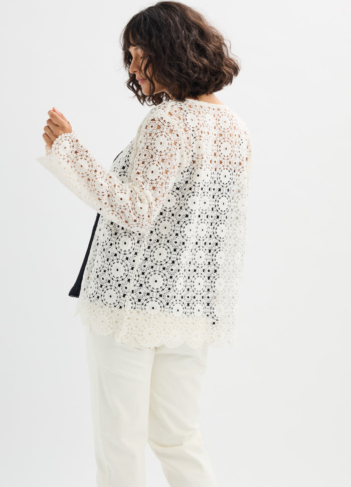 White crochet knit top with floral pattern, long sleeves, and scalloped hem, worn over black maternity tank.